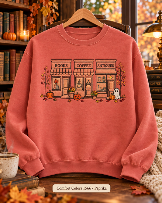 A coral pink crewneck sweatshirt hanging on a wooden hanger, featuring a graphic of a cozy storefront with 'BOOKS', 'COFFEE', and 'ANTIQUE' signs, pumpkins, a ghost, and autumn leaves. The background is a warm, cozy autumn-themed room with bookshelves, a lamp, and a mug.
